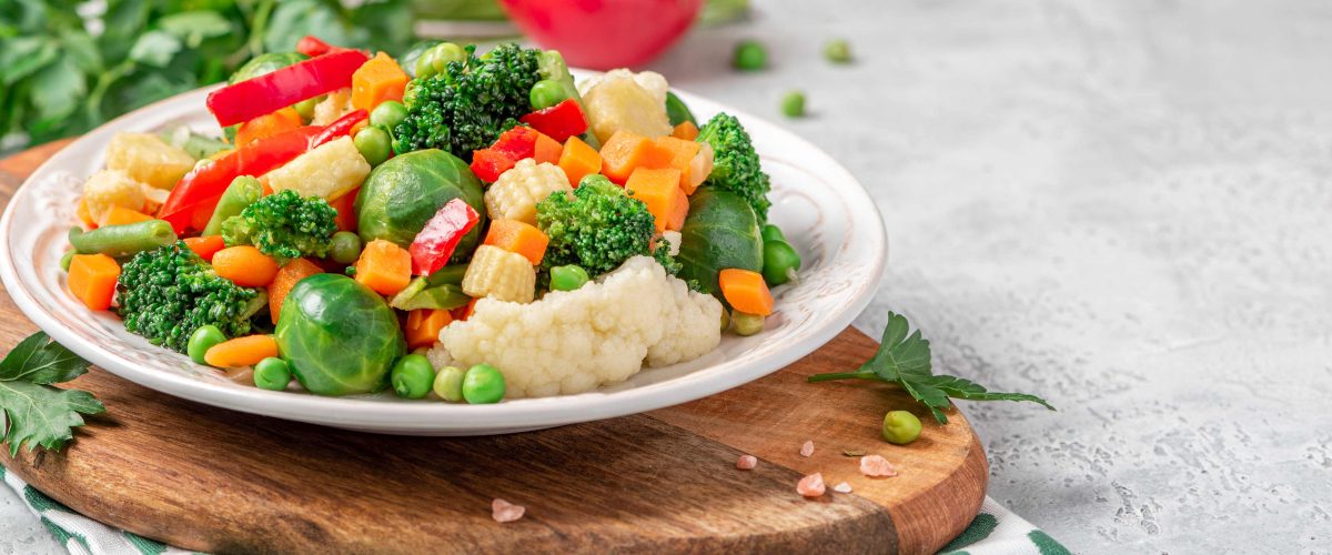 Mix of stewed vegetables in a ceramic plate on the served table. Boiled Brussels sprouts, carrots, broccoli, peas, peppers and corn. Cooked vegan or vegetarian food.